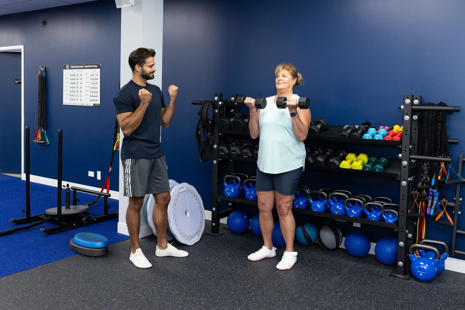 Smiling trainer and client doing dumbbell curls together beside a gym weight rack