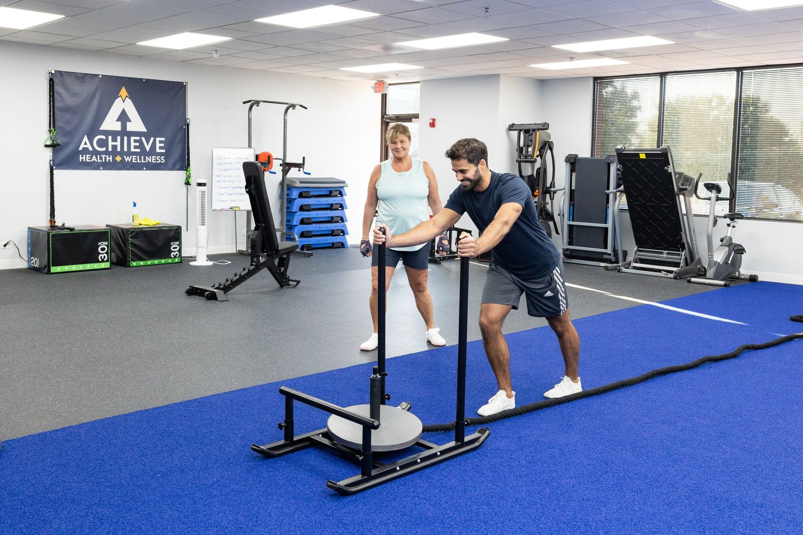 Trainer pushes a weight sled across the turf while his client watches