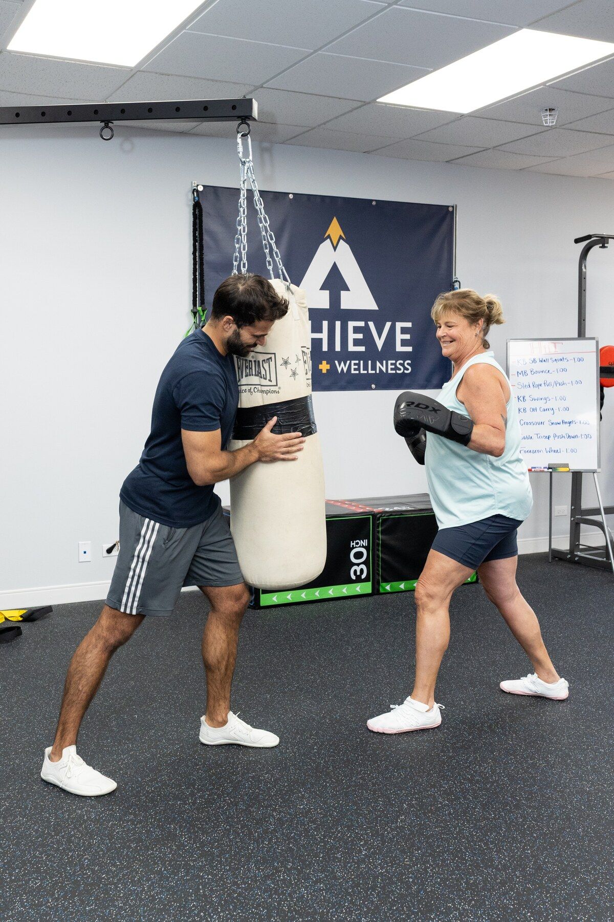 Client in boxing gloves holds a guard stance as her trainer braces the heavy bag