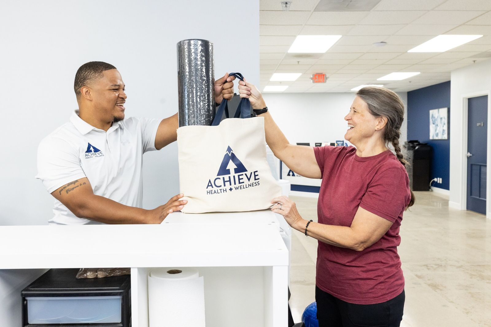 Chiropractic assistant Charles Dennis and a patient laugh as they exchange an Achieve tote bag with a foam roller at the counter