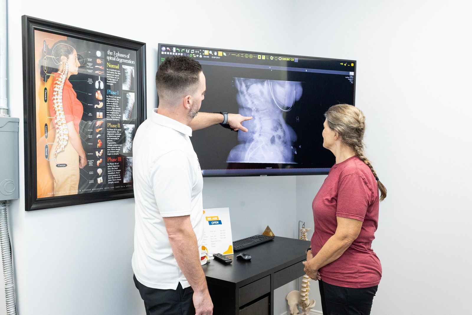 Chiropractor points to a spinal x-ray on a wall screen for a patient