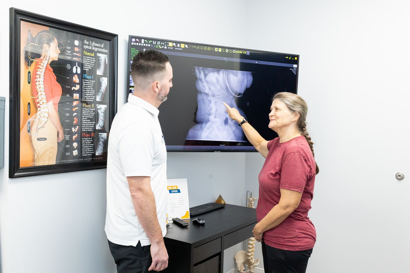 Smiling patient points to her spinal x-ray on a wall screen beside the chiropractor