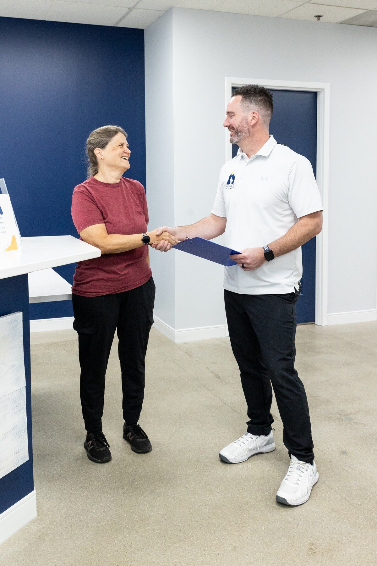 Chiropractor with a clipboard shakes hands with a smiling new patient at reception